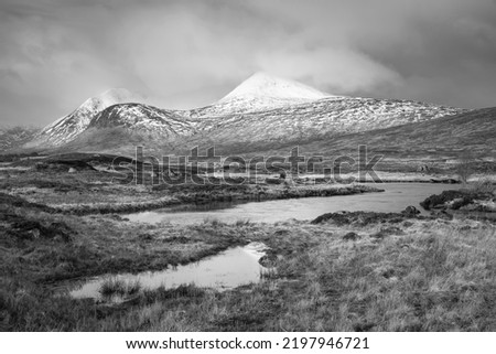Black and white Stunning Winter panorama landscape image of mountain range viewed from Loch Ba in Scottish Highlands with dramatic clouds overhead