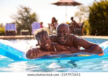 Portrait Of Smiling Senior Couple On Summer Holiday Relaxing In Swimming Pool On Inflatable