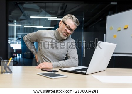 Sick senior man working inside office modern building, overtired mature businessman having severe back pain, boss massaging side to back, man at work with laptop. Royalty-Free Stock Photo #2198004241