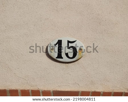 House number (address) fifteen (15) sign, black numbered white plate (closeup) against off-white wall of old building background. Grunge, texture, wallpaper