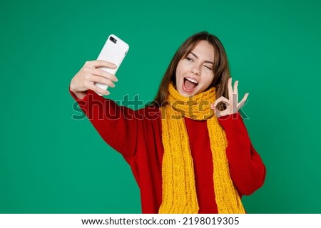 Blinking funny young brunette woman 20s in basic knitted red sweater yellow scarf doing selfie shot on mobile phone showing ok okay gesture isolated on bright green color background studio portrait