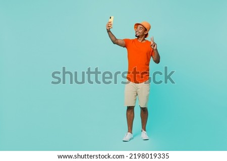 Full body young man of African American ethnicity wear orange t-shirt hat doing selfie shot on mobile cell phone post photo on social network show v-sign isolated on plain pastel light blue background