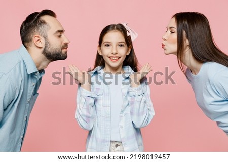 SIde view young happy parents mom dad with child kid daughter teen girl in blue clothes trying to kiss daughter isolated on plain pastel light pink background. Family day parenthood childhood concept