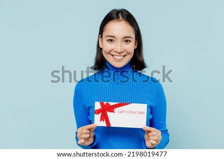 Fascinating fun smiling young woman of Asian ethnicity 20s years old wears blue shirt hold gift certificate coupon voucher card for store isolated on plain pastel light blue background studio portrait