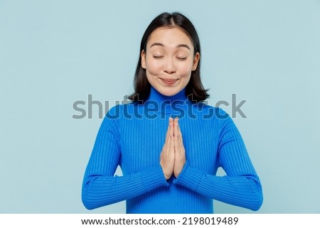 Young woman of Asian ethnicity 20s years old wears blue shirt hands folded in prayer gesture begging about something keeps eyes closed isolated on plain pastel light blue background studio portrait Royalty-Free Stock Photo #2198019489