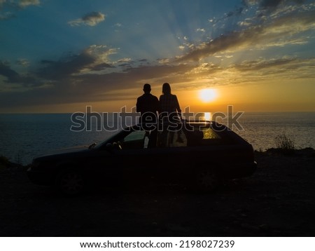 Silhouette of a happy couple sitting on the roof of a car on a cliff in front of the sea with a beautiful orange-blue sky at sunset, view from a drone. Concept of vacations and travel, romance
