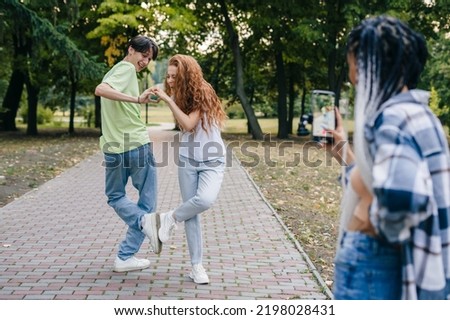 Cheerful beautiful girl standing in the park with a camera and smiling while recording a video of her two friends dancing while showing the heart sign