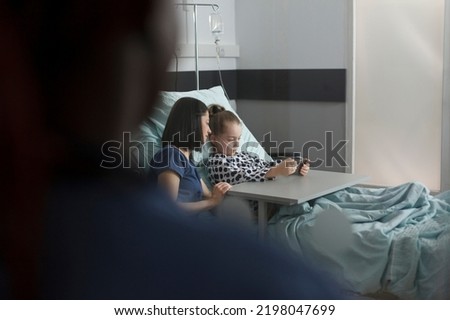 Caring parent sitting beside hospitalized sick daughter watching cartoons on smartphone. Ill little girl playing games on mobile phone while resting in hospital pediatrics ward patient bed.