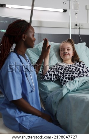 Childcare healthcare facility staff high fiving ill kid under medical treatment. Nurse doing high five gesture with sick girl resting in hospital pediatric ward patient bed. Royalty-Free Stock Photo #2198047727