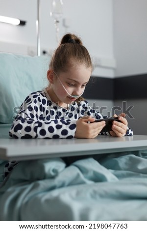 Hospitalized ill kid watching cartoons on phone while sitting inside pediatric healthcare clinic ward. Pretty little sick girl resting on bed while playing games on modern smartphone.