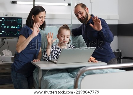 Sick little girl sitting with parents on patient bed while waving at laptop video call inside hospital pediatrics ward. Ill kid in healthcare clinic talking with relatives on virtual online call.