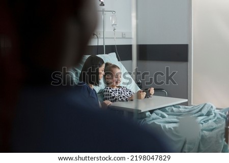 Mother sitting beside hospitalized daughter under treatment while watching cartoons on smartphone. Ill little girl resting in hospital pediatrics ward while playing games on modern mobile phone Royalty-Free Stock Photo #2198047829