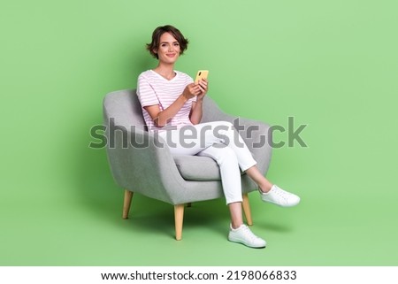 Full size photo of cute young woman sit armchair relaxing hold device typing dressed trendy striped look isolated on green color background