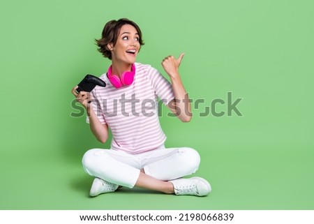 Full length photo of nice young girl point empty space playstation earphones dressed trendy striped look isolated on green color background