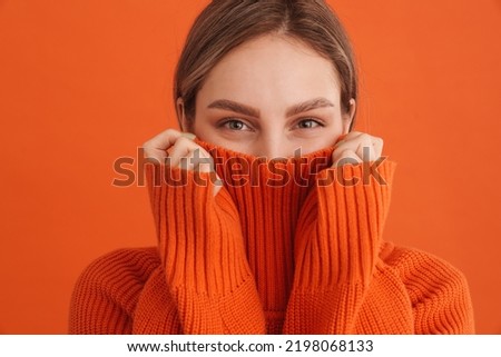 Young cute girl hiding in sweaters neck over isolated orange background
