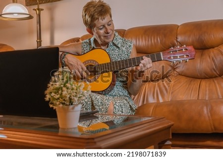 Adult woman learning to play guitar at home by video call. Horizontal photography.