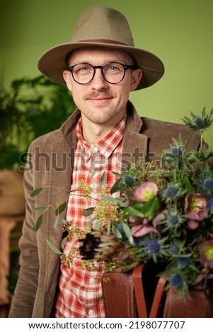 Poet or writer portrait. Attractive male person in eyeglasses and hat staying with flower bouquet and looking in camera in flower shop. High quality vertical image