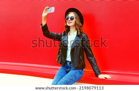 Portrait of beautiful happy woman taking selfie with smartphone wearing black round hat on red background