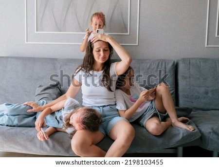 Tired young mother sits on couch and holds her head while her three naughty daughters play games on smartphones. lifestyle family together portrait. Royalty-Free Stock Photo #2198103455