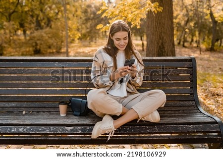 candid attractive young smiling woman sitting on bench in autumn park using phone wearing checkered coat, happy mood, fashion style trend Royalty-Free Stock Photo #2198106929