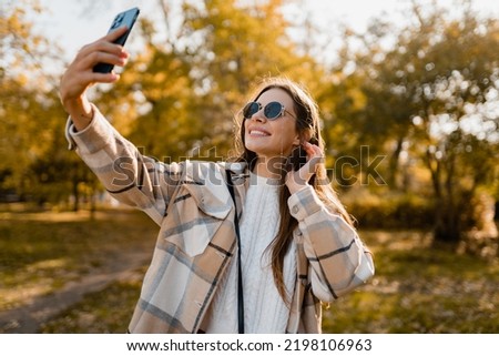 attractive young smiling woman walking in autumn park taking selfie pictures using smartphone, wearing checkered coat, sunglasses, happy mood, fashion style trend