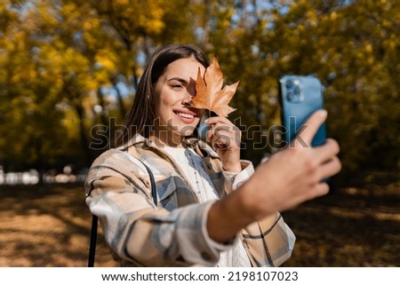 attractive young smiling woman walking in autumn park taking selfie pictures with leaf using smartphone, wearing checkered coat, happy mood, fashion style trend