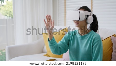 Asian people girl fun play smart live VR 360 hand touch in metaverse app look at augmented AR VFX game platform on goggles glasses headset sit at sofa couch enjoy leisure lifestyle vacation at home.