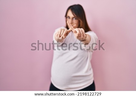Pregnant woman standing over pink background rejection expression crossing fingers doing negative sign 
