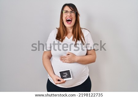 Pregnant woman holding baby ecography angry and mad screaming frustrated and furious, shouting with anger looking up. 