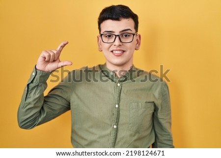 Non binary person standing over yellow background smiling and confident gesturing with hand doing small size sign with fingers looking and the camera. measure concept. 