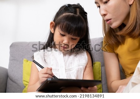Young Asian girl using digital tablet for painting picture or study online lesson at home, with mother support and teaching the lesson inside of living room. Online education or home school concept.