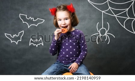 Kid in Halloween costume horns having fun, adorable happy child smiles and eats hallowen pumpkin cookie, studio portrait of little girl with drawing bats and spider. Trick or treat, Halloween theme.