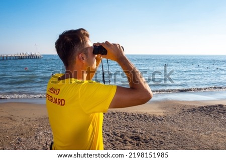 Lifeguard on the beach looking through binoculars. Safety while swimming, handsome brunette male lifeguard on the beach,back shot looking at the blue sea, copy space. Royalty-Free Stock Photo #2198151985
