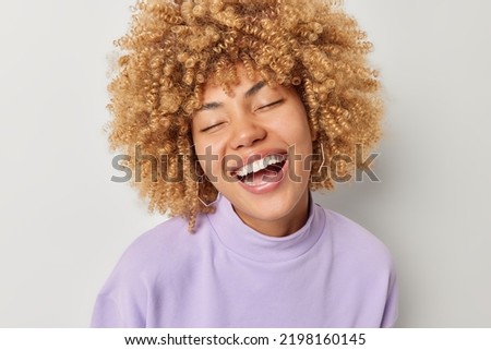 Portrait of happy positive woman keeps eyes closed smiles broadly shows white teeth dressed in purple pullover enjoys life feels very glad isolated over grey studio background. People and emotions Royalty-Free Stock Photo #2198160145