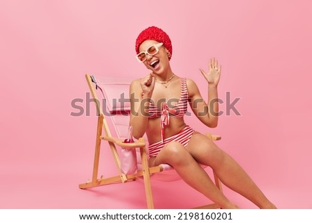 Beautiful woman wears hat striped swimwear and sunglasses has good mood keeps hand near mouth as if microphone sings song keeps palm raised poses on deck chair enjoys summer holidays at beach