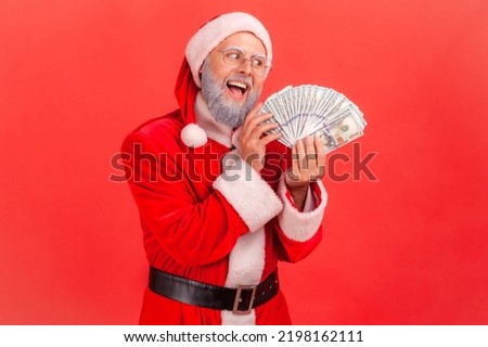 Portrait of elderly man with gray beard wearing santa claus costume with excited expression, greedy male with fan of dollars, looking at banknotes. Indoor studio shot isolated on red background.