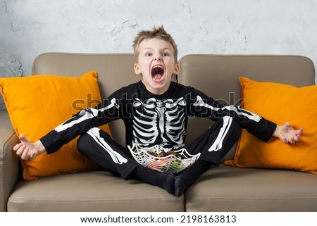 Happy shocked crazy shouts halloween boy having fun at home eat scary candy and looking at camera wearing Halloween costume skeleton on home background. Royalty-Free Stock Photo #2198163813