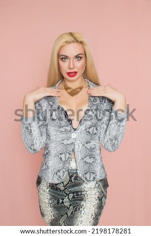 Fashion photo of a beautiful elegant young woman in pretty snake suit, jacket blazer, top, skirt, massive chain around the neck posing on pink background. Studio shot.