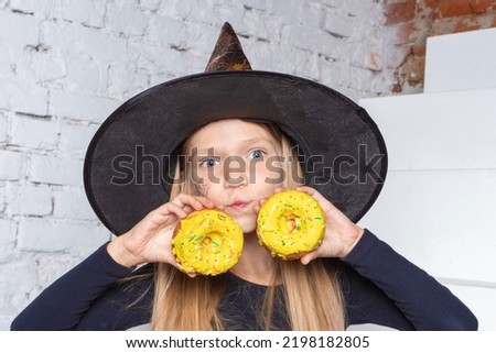 Halloween kids. A smiling little blonde girl in a witch costume holding yellow donuts in her hands, sitting on the steps of the house. Happy Halloween