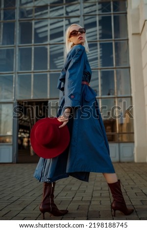 Fashionable confident blonde woman wearing trendy sunglasses, blue midi trench coat, belt, leather ankle boots, holding stylish hat. Female fashion conception. Outdoor full-length portrait Royalty-Free Stock Photo #2198188745