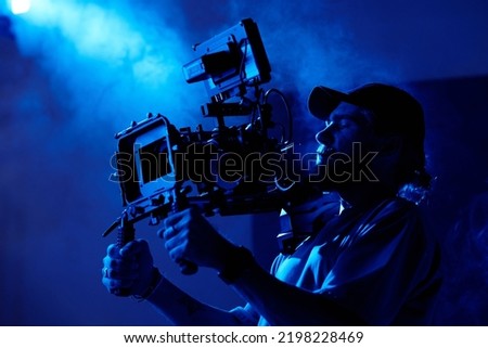Young videographer in casualwear shooting commercial video in dark room or studio full of smoke lit by dark blue light Royalty-Free Stock Photo #2198228469