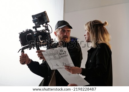 Young bearded cameraman and his assistant with documents consulting about shooting while standing in front of camera in studio Royalty-Free Stock Photo #2198228487