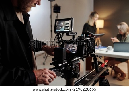 Young man with videocamera standing in studio in front of his assistant with documents and female model taking part in commercial shooting Royalty-Free Stock Photo #2198228665