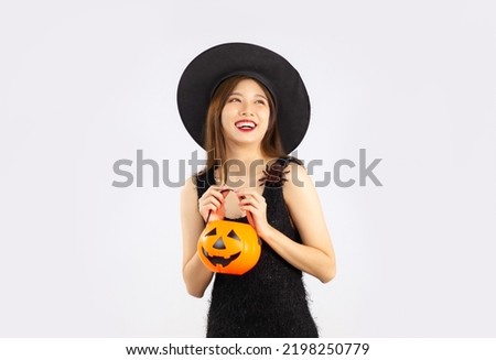 Halloween theme young asian woman in black costume wearing witch hat carrying pumpkin lantern on white background.