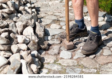a gardener in dirty work clothes and boots holds a two-bladed iron hammer 