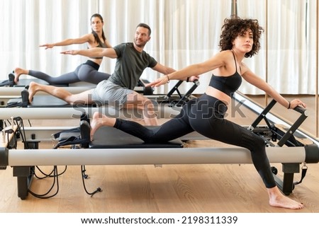 Group of barefoot women and man in activewear spreading arms and looking away while doing stretching exercise on reformers during pilates training in gym Royalty-Free Stock Photo #2198311339