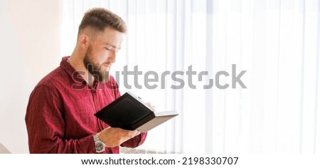 Business bearded man stands against window in office and writes down plans in a notebook in a dark burgundy shirt. Concept banner