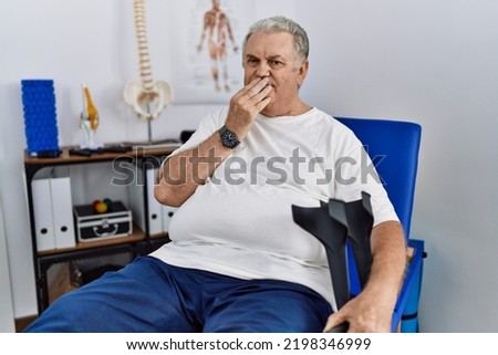 Senior caucasian man at physiotherapy clinic holding crutches laughing and embarrassed giggle covering mouth with hands, gossip and scandal concept  Royalty-Free Stock Photo #2198346999