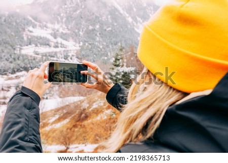 Rear view portrait of young blond woman taking a picture with smartphone at snow mountain. Wears black jacket and yellow hat for cold weather at winter time.