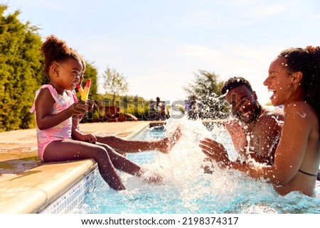 Family On Summer Holiday With Two Girls Eating Ice Lollies By Swimming Pool Splashing Parents Royalty-Free Stock Photo #2198374317